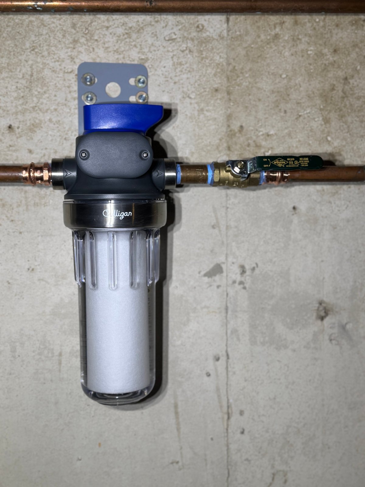 Water filter system with transparent canister and blue cap mounted on copper tubing against concrete wall