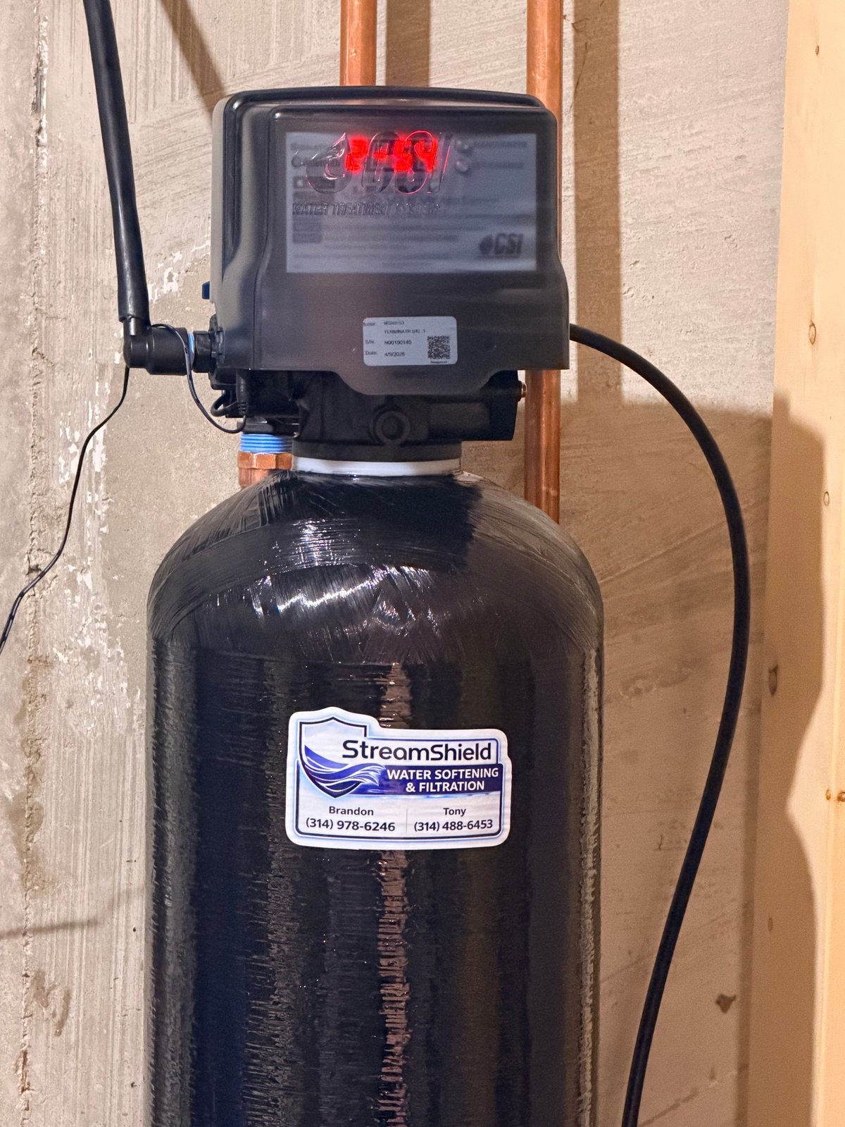 StreamShield water softener system with digital control head mounted on black resin tank against wall with copper pipes