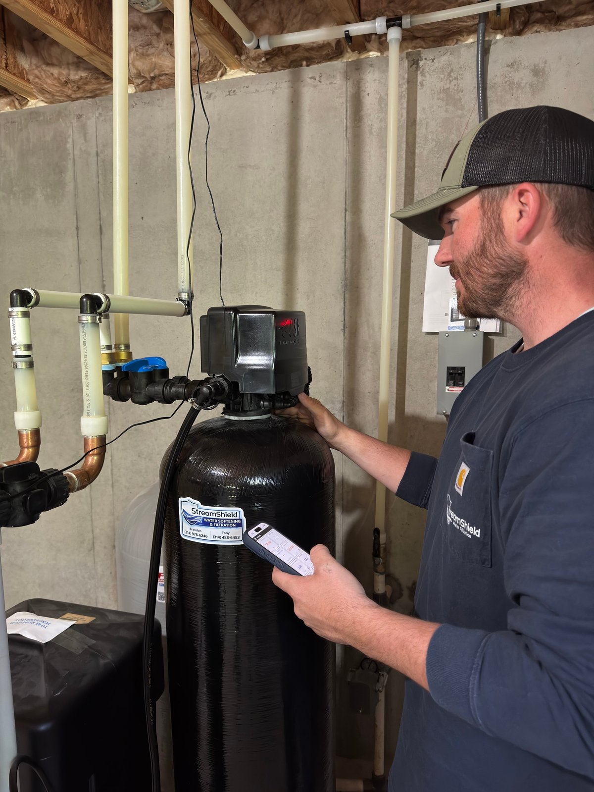Technician in black cap and shirt checking readings on a water softener system in basement utility area